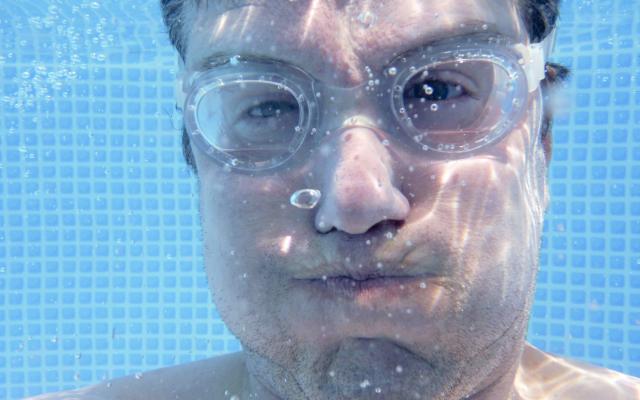 Gezicht van een man onderwater. Hij draagt een duikbril en heeft zijn wangen opgeblazen.